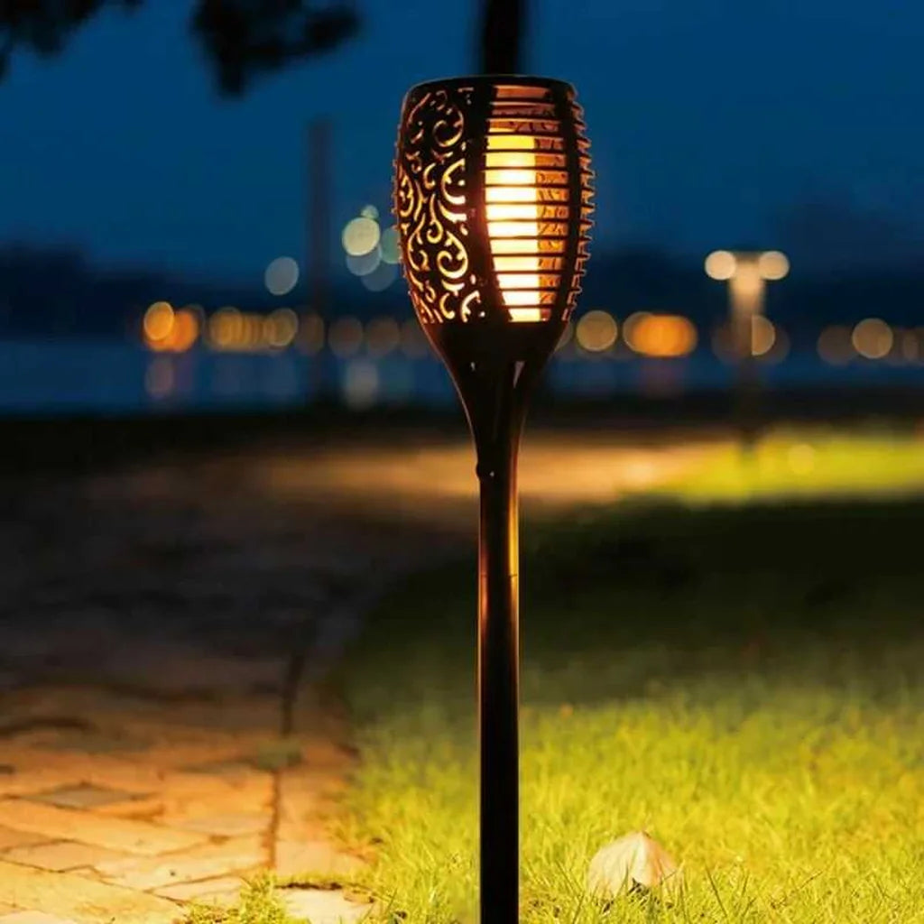 Antorcha solar LED con efecto de fuego iluminando sendero de jardín durante la noche.