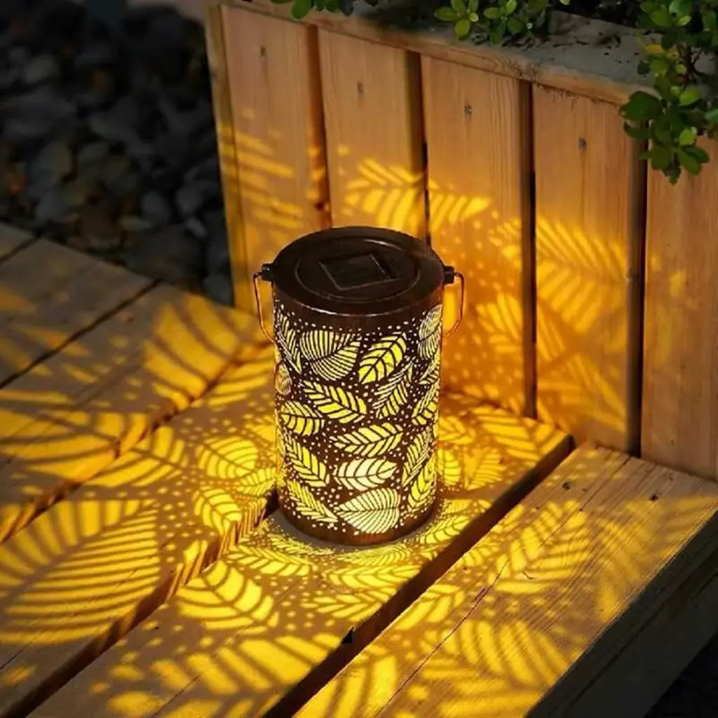 Farol solar decorativo proyectando sombras de hojas en una terraza de madera, ideal para iluminar jardines con diseño cálido y natural.
