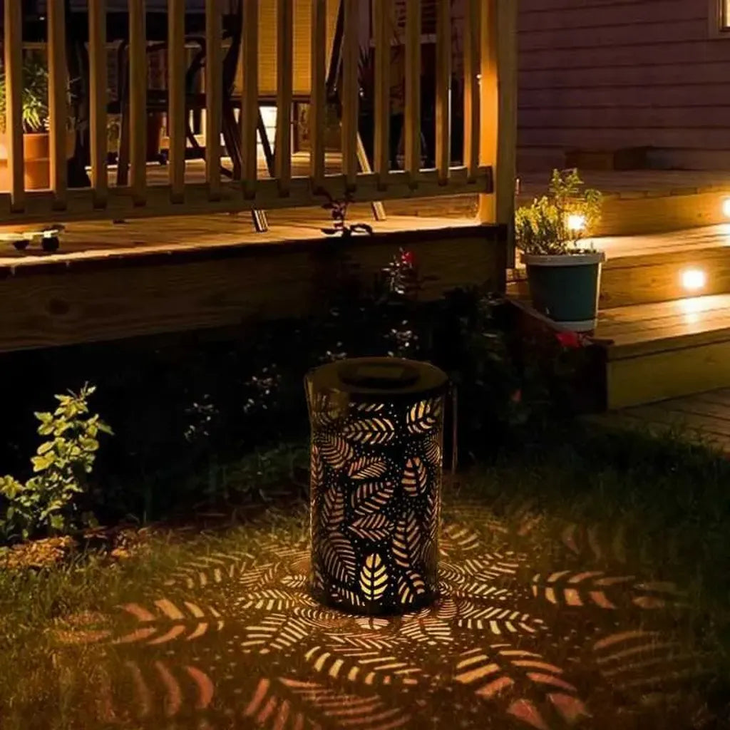 Farol solar decorativo iluminando jardín de noche, proyectando sombras artísticas sobre el césped junto a una terraza de madera.