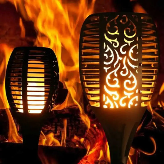 Antorchas solares LED con efecto llama encendidas frente a fondo de fuego real, diseño decorativo para jardín o terraza