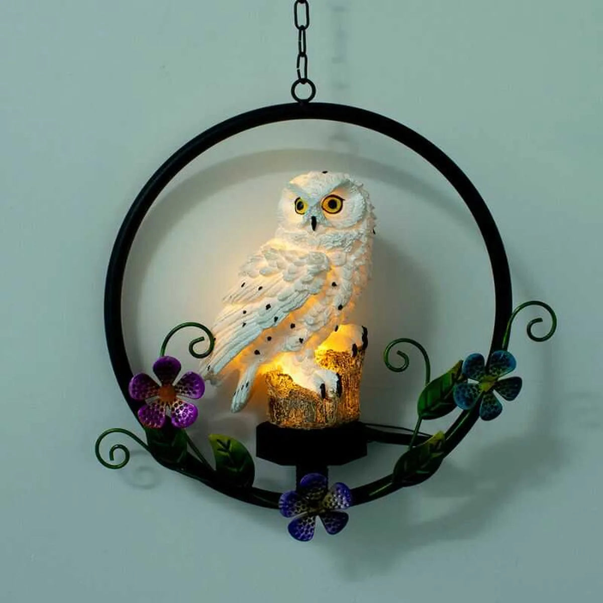 Lámpara solar de búho iluminado colgando en pared con aro metálico y flores decorativas, ideal para interior o exterior.