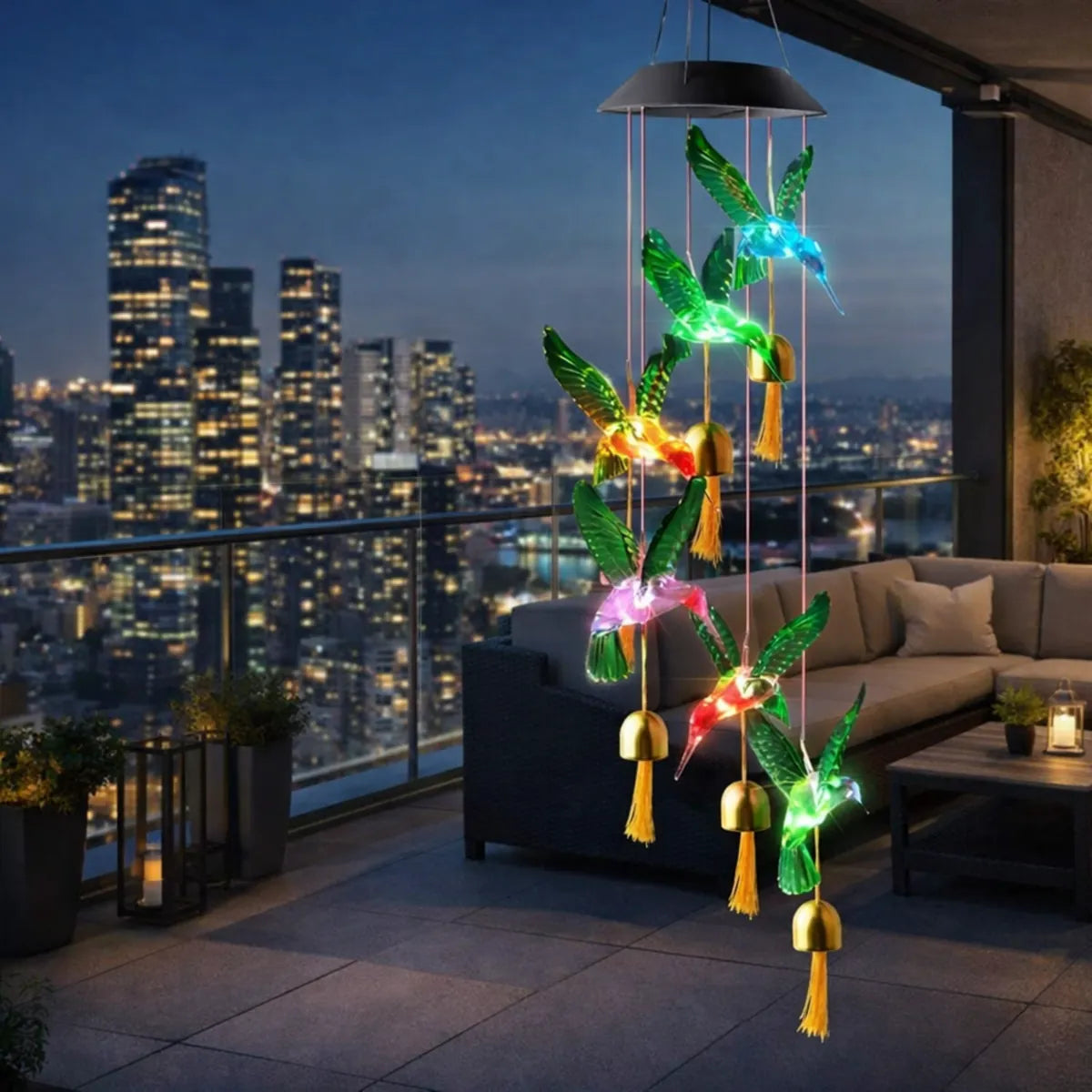 Colgante solar de colibríes LED multicolor en terraza de edificio con vista nocturna, ideal para decoración de balcón o exterior.