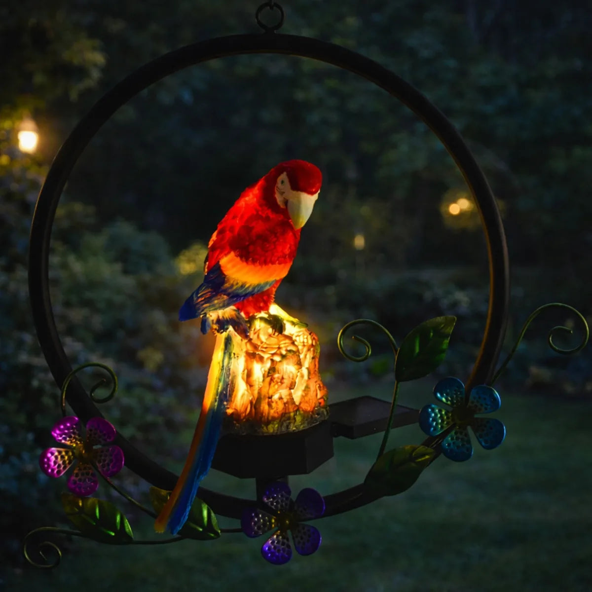 Lámpara solar decorativa de loro iluminado en jardín nocturno con aro metálico y flores, ideal para exterior.
