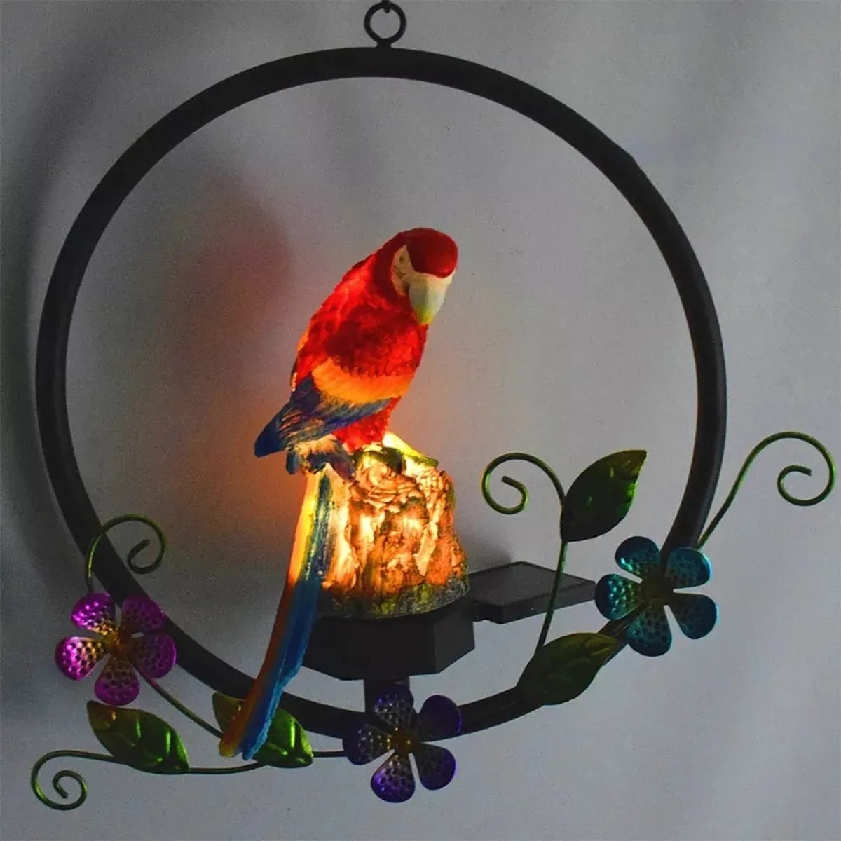 Lámpara solar decorativa con figura de loro iluminado sobre aro metálico con flores, ideal para jardín o terraza.